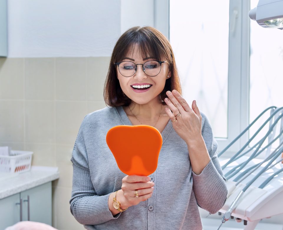 Happy Woman Dentist Patient With Mirror In Hands Looking At Her Teeth