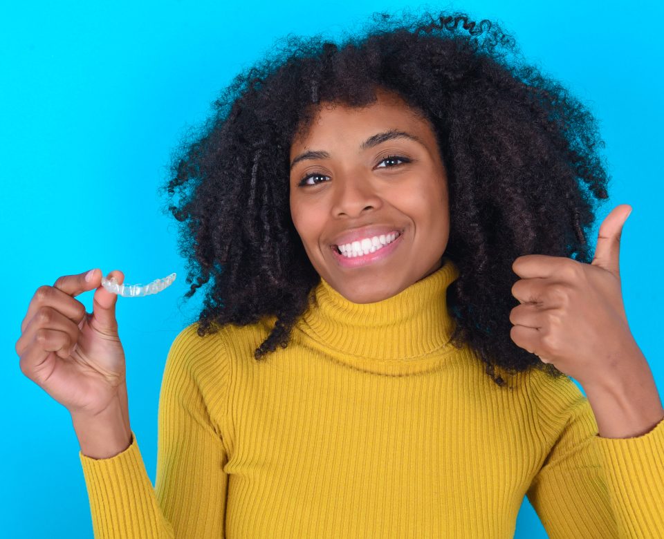 Young Woman With Afro Hairstyle Wearing Yellow Turtleneck Over Blue Background Holding An Invisible Braces Aligner And Rising Thumb Up, Recommending This New Treatment. Dental Healthcare Concept.