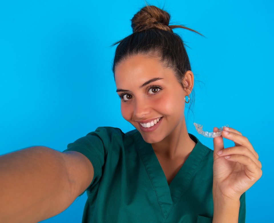 Beautiful Doctor Woman Wearing Medical Uniform Over Blue Background Make Selfie Holding An Invisible Braces Aligner, Recommending This New Treatment. Dental Healthcare Concept.