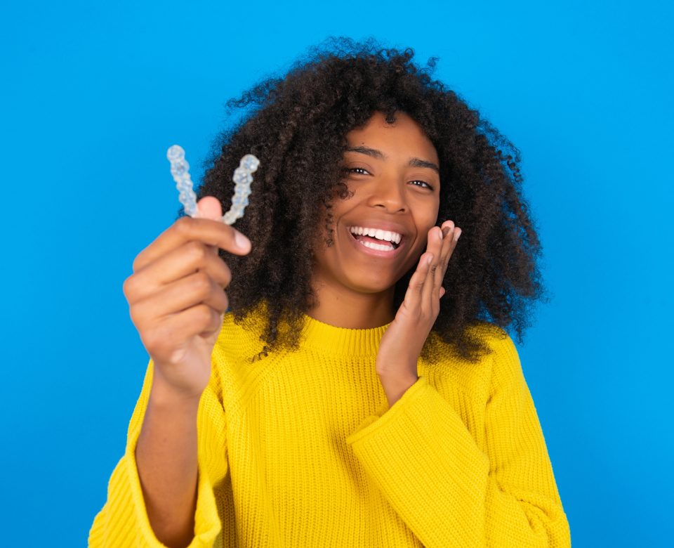 Happy Young Woman With Afro Hairstyle Wearing Orange Crop Top Over Blue Wall Holding And Showing At Camera An Invisible Aligner While Laughing. Dental Healthcare And Confidence Concept.